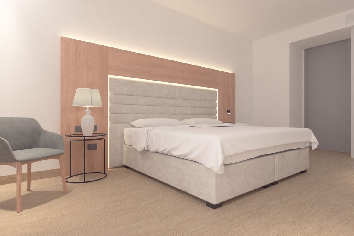 A modern hotel room with a large bed, a bedside table with a lamp, a chair, and a wardrobe.