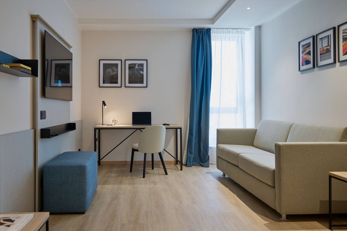 Una sala moderna con un sofá, un escritorio, una silla y un reposapiés azul. Hay cuadros enmarcados en la pared y una ventana con cortinas azules.