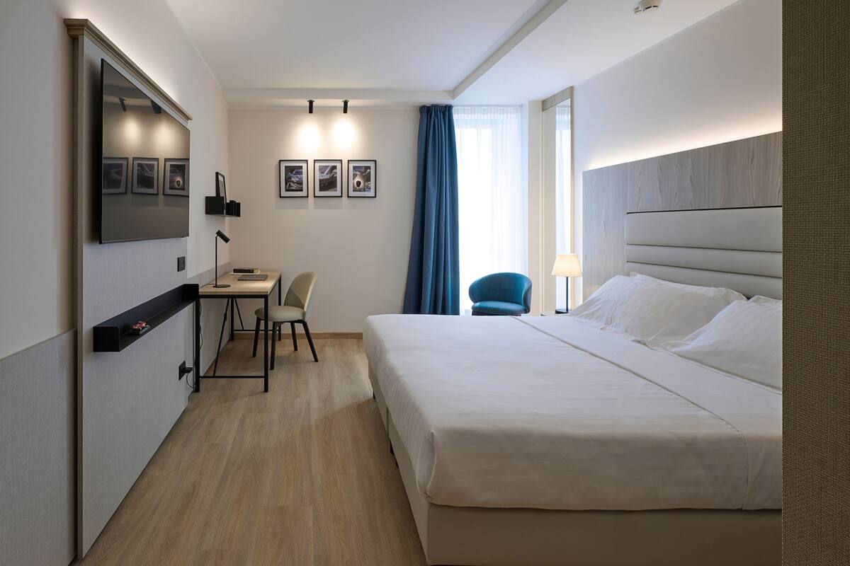 Una habitación de hotel con una cama grande, un escritorio con silla, un televisor montado en la pared y una ventana con cortinas.