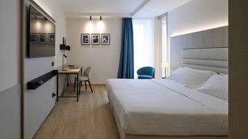 A hotel room with a large bed, a desk with a chair, a TV mounted on the wall, and a window with curtains.