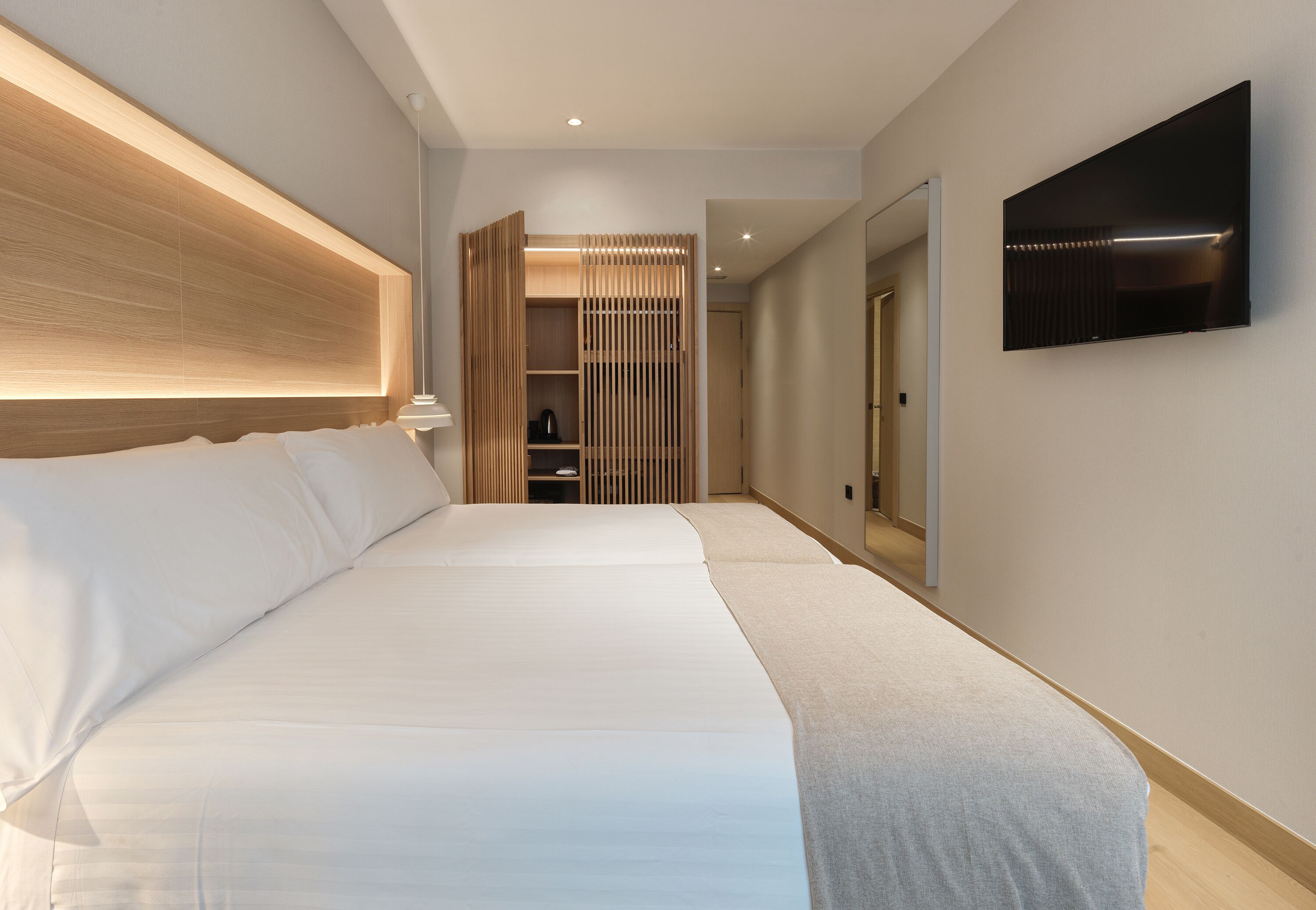 A modern hotel room with a large bed, a walk-in closet, a TV mounted on the wall, and a wooden headboard.