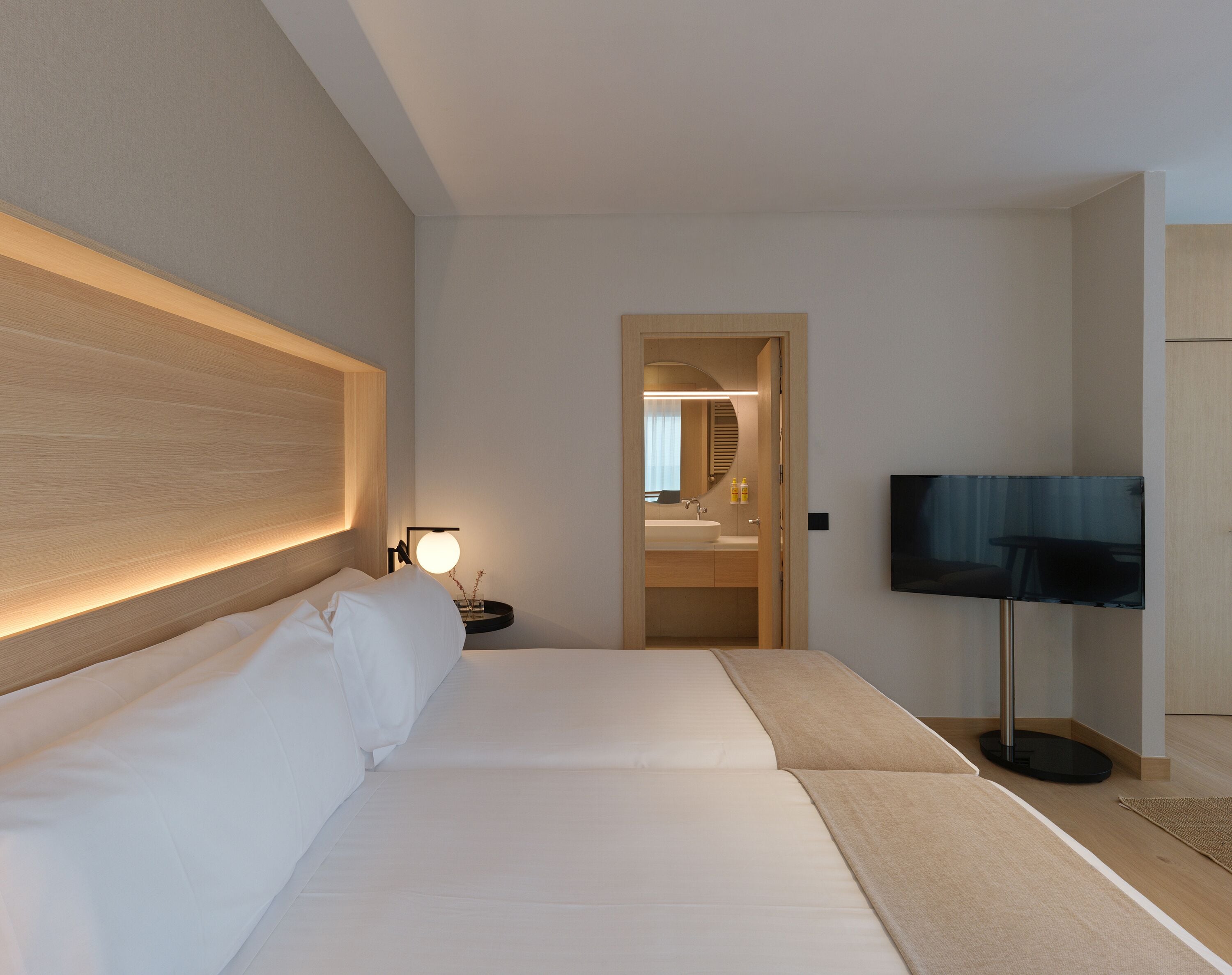 A hotel room with a large bed, a bedside table with a lamp, a TV, and a bathroom visible through an open door.