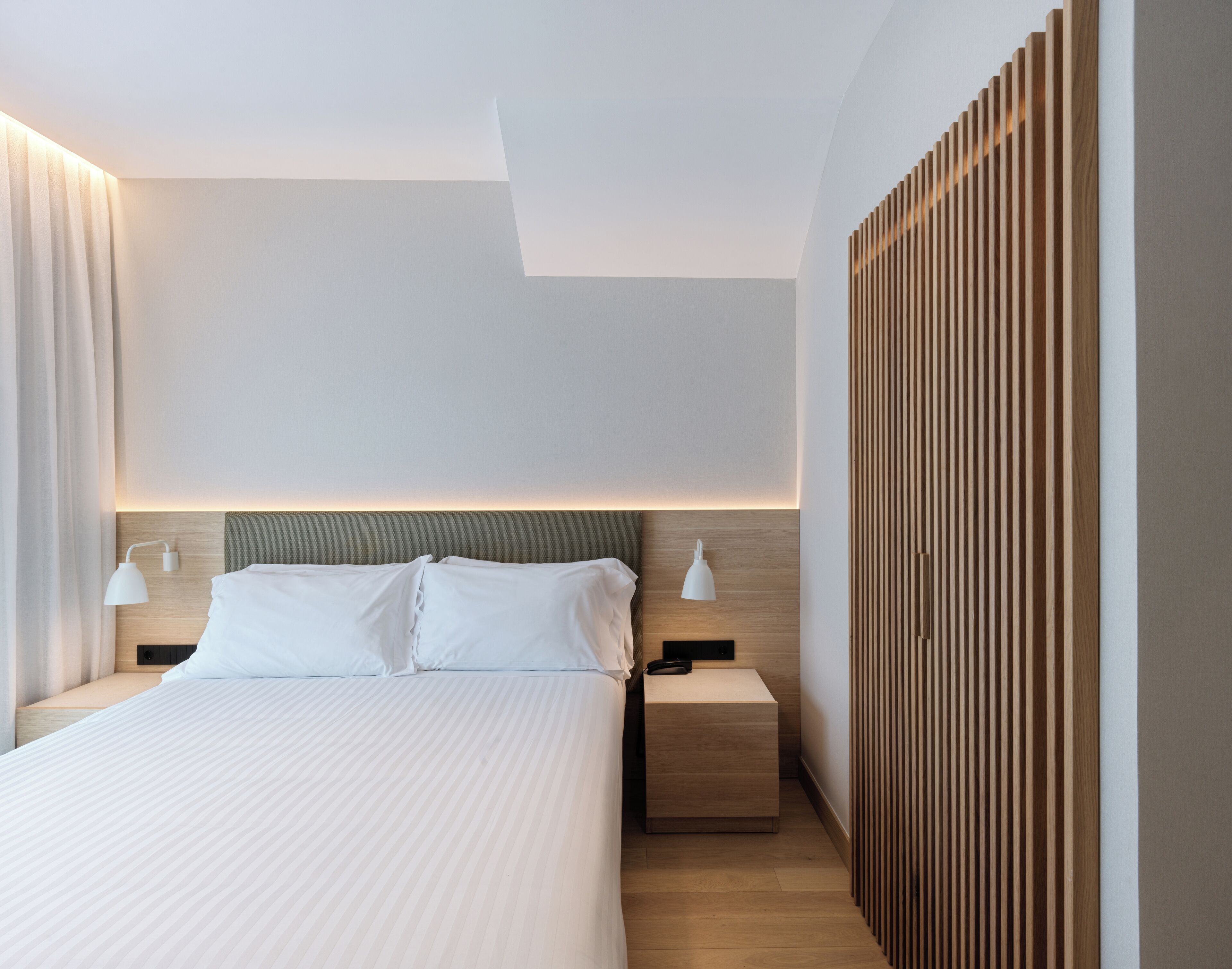 A modern hotel room with a large bed, two bedside tables with lamps, a wooden headboard, and a built-in wooden panel on the wall.