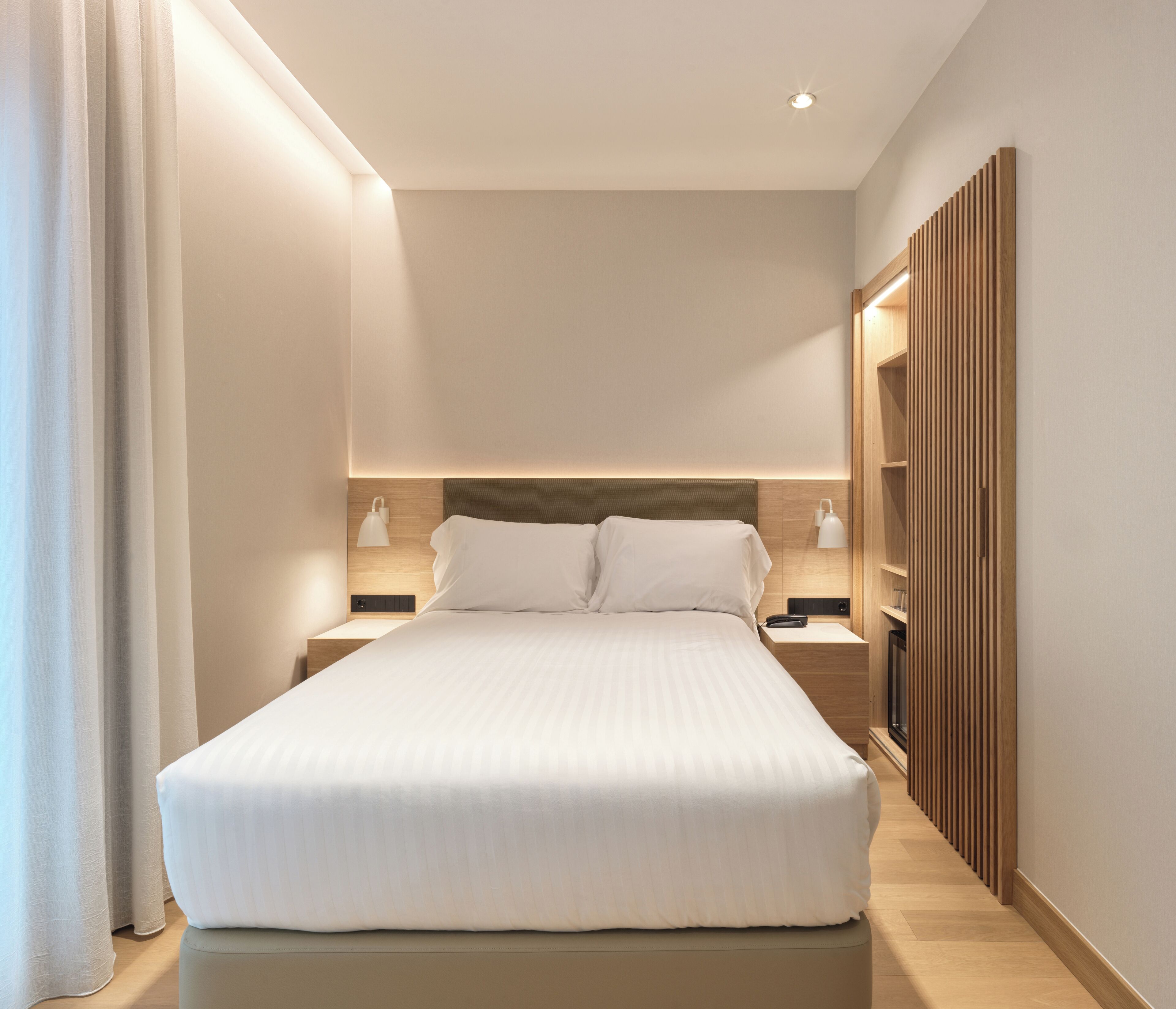 A modern hotel room with a large bed, bedside tables, and a wardrobe.