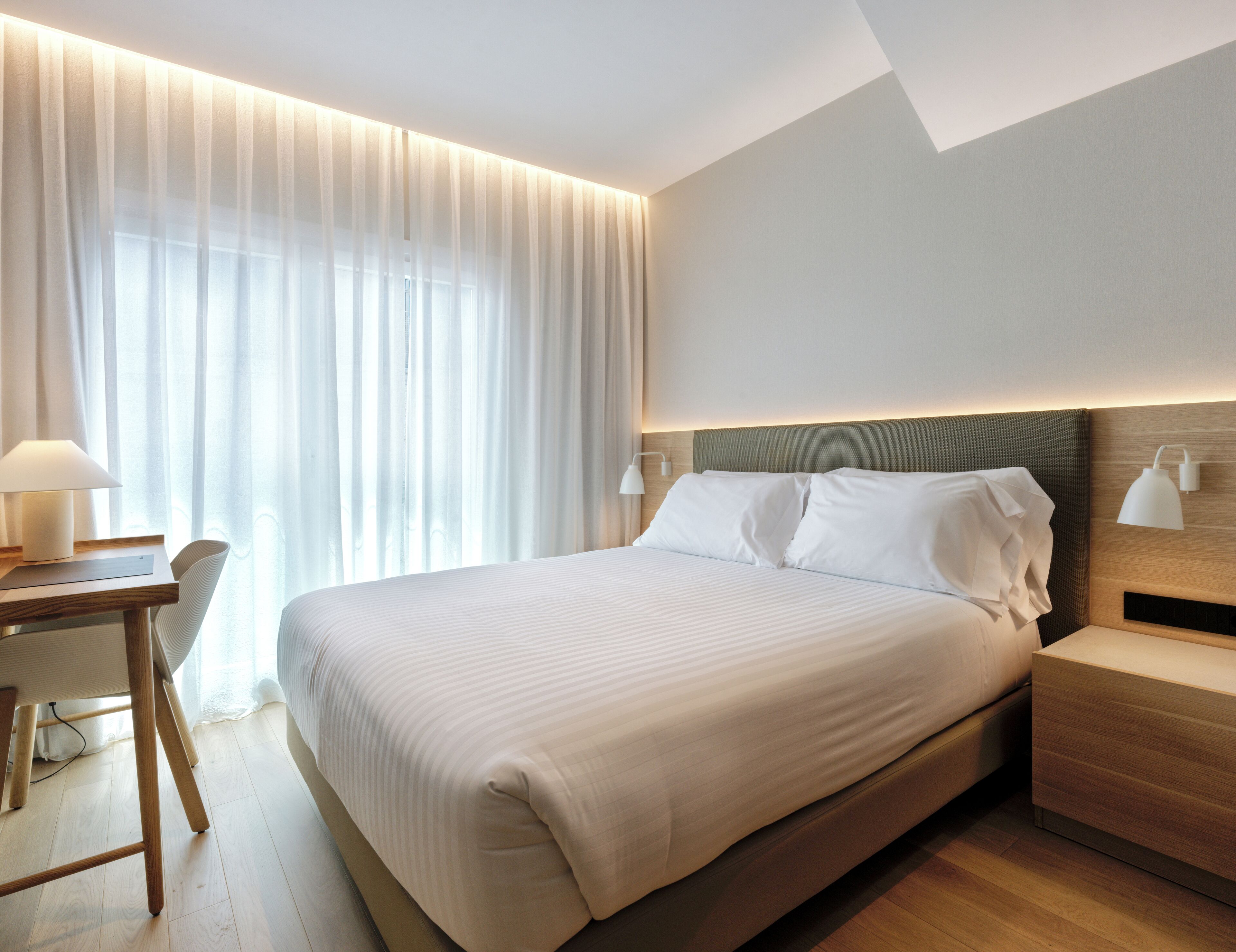 A hotel room with a large bed, bedside tables, a desk with a chair, and sheer curtains.