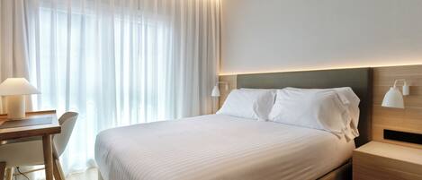 A hotel room with a large bed, bedside tables, a desk with a chair, and sheer curtains.
