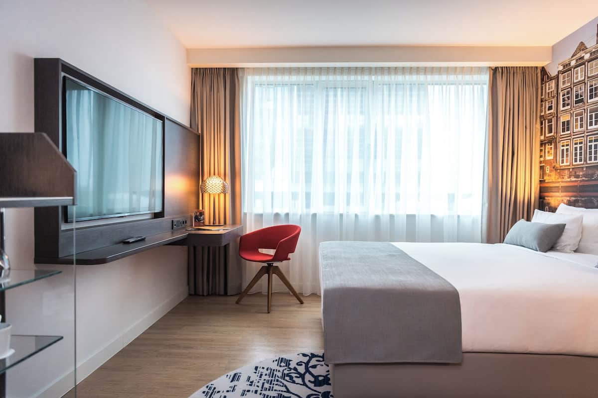 A modern hotel room with a large bed, a desk with a flat-screen TV, a red chair, and a large window with curtains.