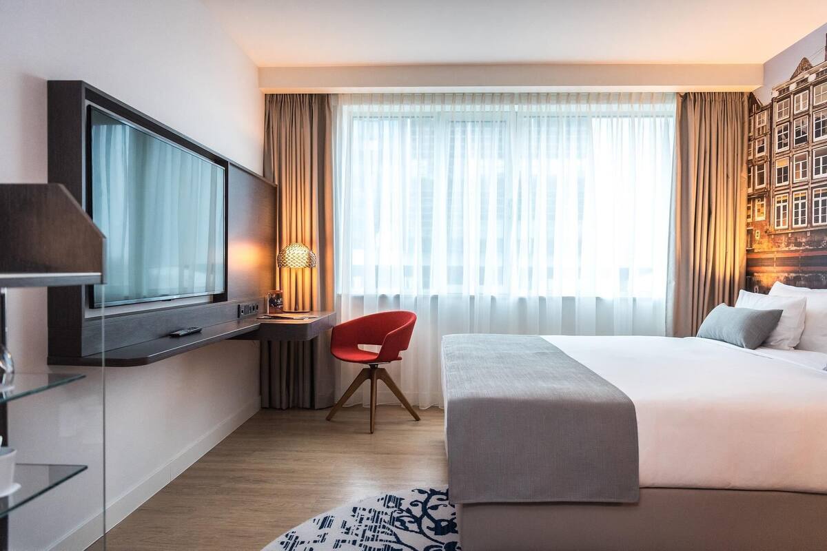 Una habitación de hotel con cama, escritorio con silla, televisor y un ventanal con cortinas.