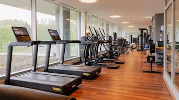 A gym with treadmills, ellipticals, and weight machines, large windows, and a wooden floor.