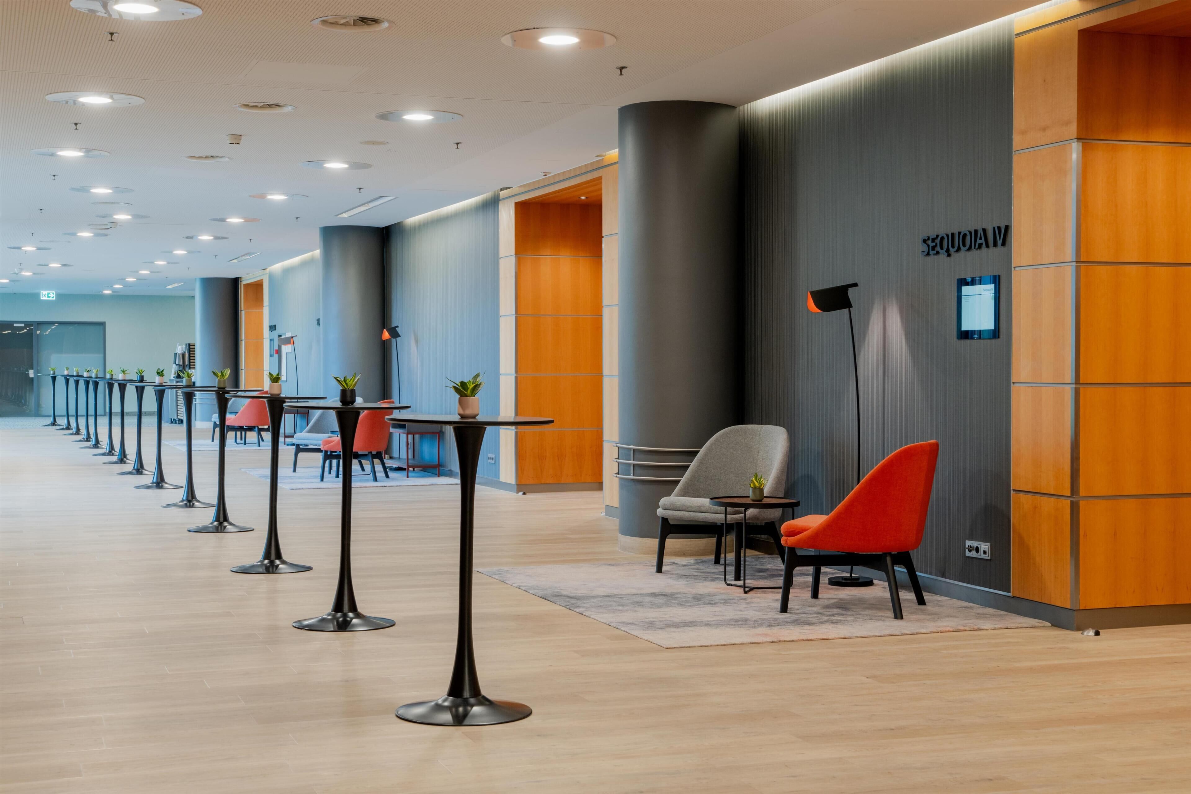 A modern hotel lobby with a row of tables and chairs, a wooden accent wall, and a sign indicating room number IV.