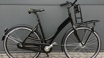 A stationary black bicycle with a front basket and a rear rack, parked against a grey wall.