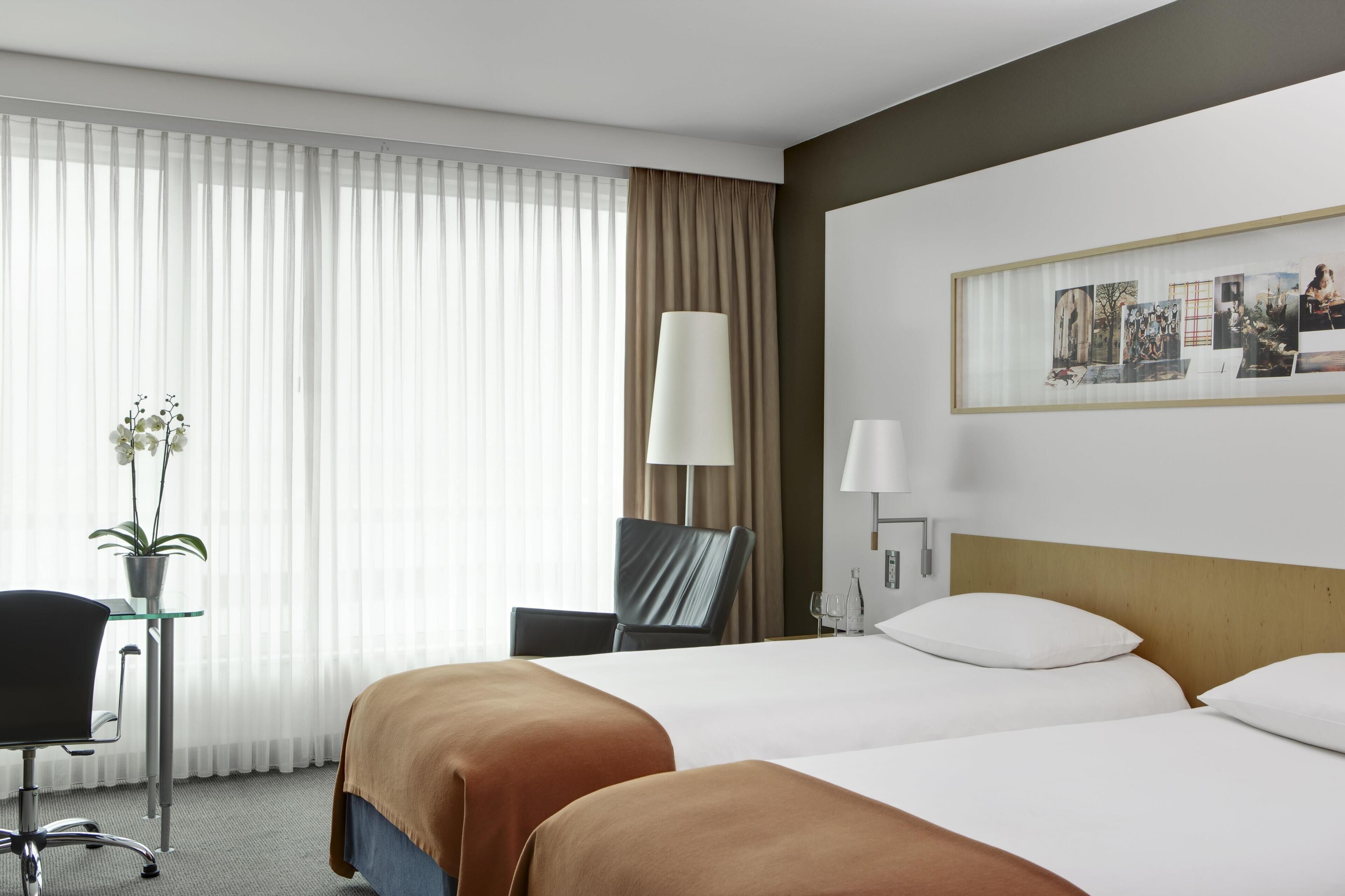 A hotel room with two beds, a desk with a chair, a lamp, a framed picture on the wall, and a window with curtains.