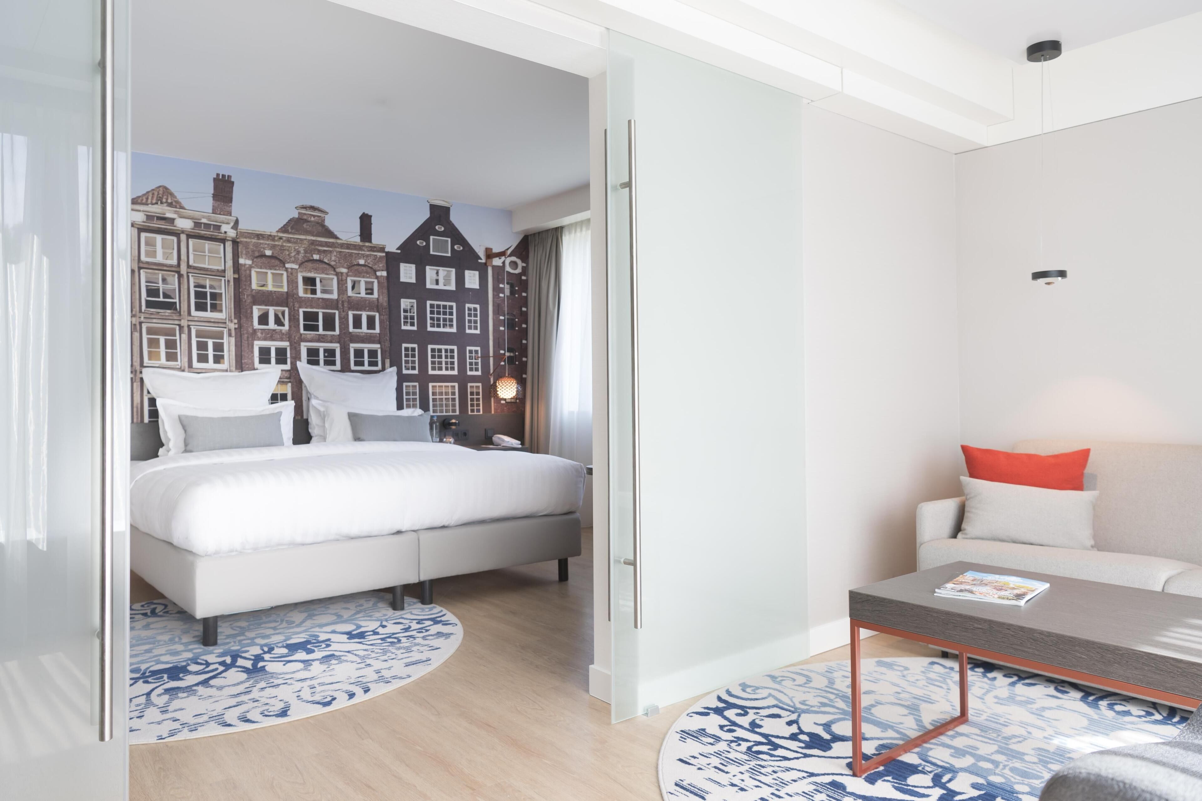 Una habitación de hotel moderna con una cama, un sofá, una mesita de centro y vistas a un paisaje urbano a través de un ventanal.