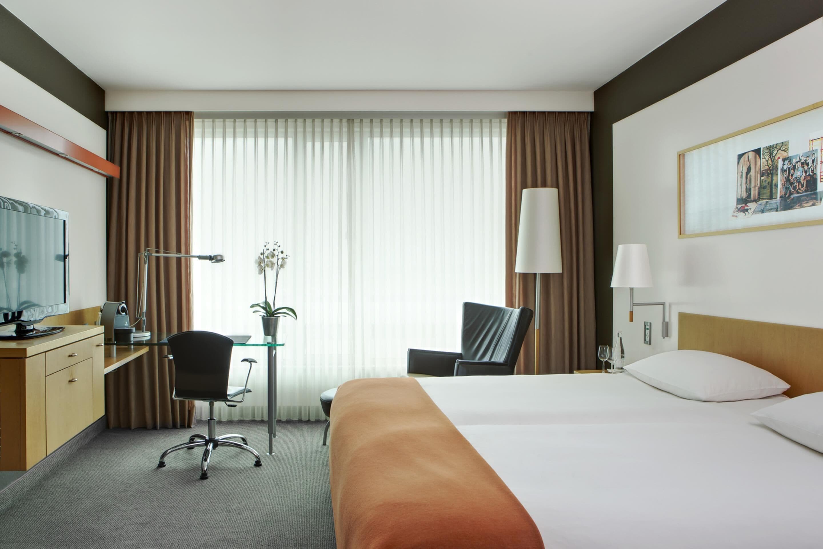 A hotel room with a bed, desk, chair, lamp, and a window with curtains.