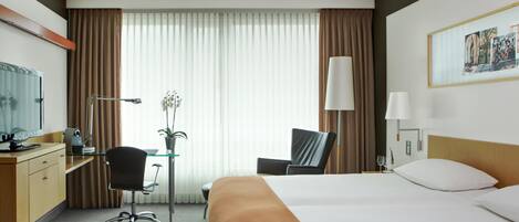 Une chambre d’hôtel avec un lit, un bureau, une chaise, une lampe et une fenêtre avec des rideaux.