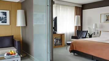 Et hotelværelse med en seng, et skrivebord med computer, en stol, en sofa og en glasvæg.