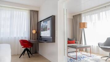 Una habitación de hotel moderna con un televisor de pantalla plana montado en la pared, una zona de comedor con mesa y sillas, y un ventanal con cortinas translúcidas.