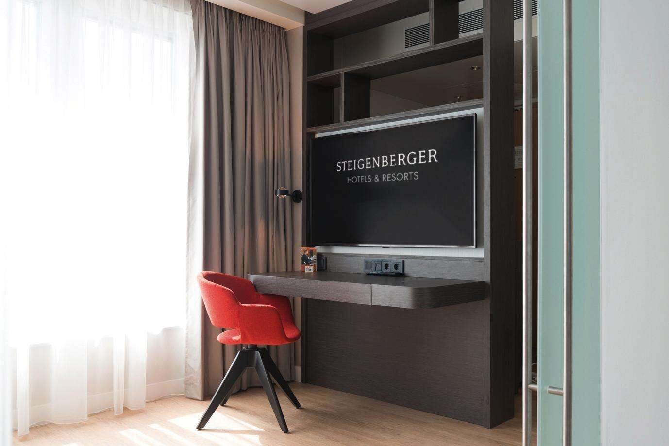 A modern hotel room with a dark wood desk, a red chair, and a large TV screen displaying 'Steigenberger Hotels & Resorts'.