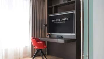 A modern hotel room with a dark wood desk, a red chair, and a large TV screen displaying 'Steigenberger Hotels & Resorts'.