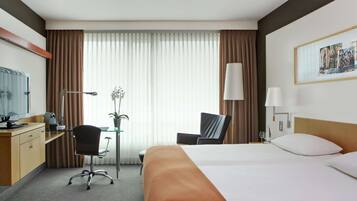 A hotel room with a bed, desk, chair, and a large window with curtains.