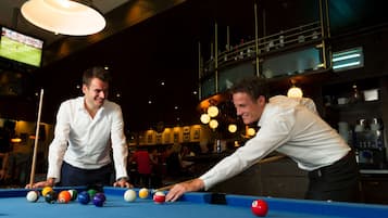 Twee personen spelen biljart in een ruimte met een bar en een televisiescherm waarop een sportevenement te zien is.