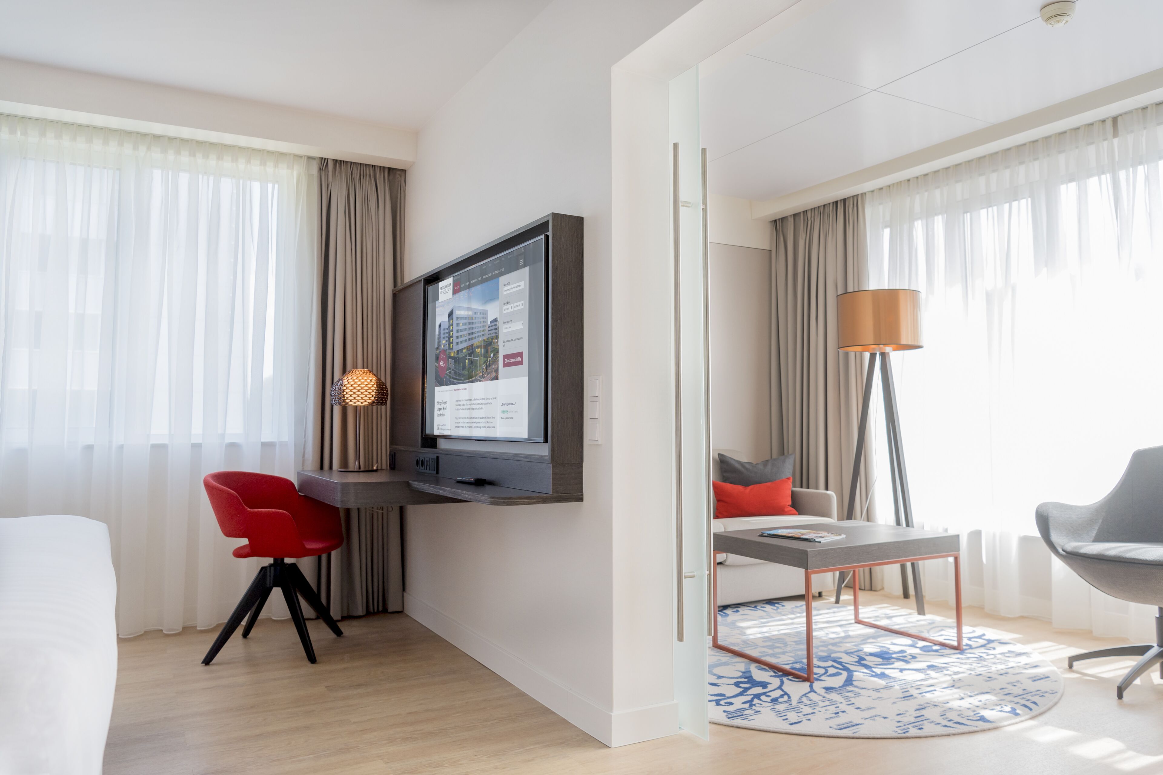 Una habitación de hotel moderna con una televisión de pantalla plana montada en la pared, un comedor con mesa y sillas, y un ventanal con cortinas translúcidas.