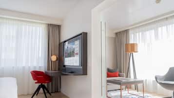 Een moderne hotelkamer met een wandgemonteerde flatscreen-tv, een zithoek met een tafel en stoelen, en een groot raam met dunne gordijnen.