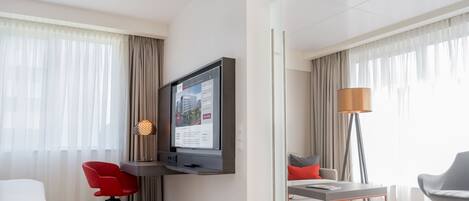 Ein modernes Hotelzimmer mit einem an der Wand befestigten Flachbildfernseher, einem Essbereich mit Tisch und Stühlen sowie einem großen Fenster mit durchsichtigen Vorhängen.