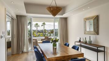 A dining area with a wooden table, blue chairs, a hanging light fixture, and a view of palm trees through large windows.