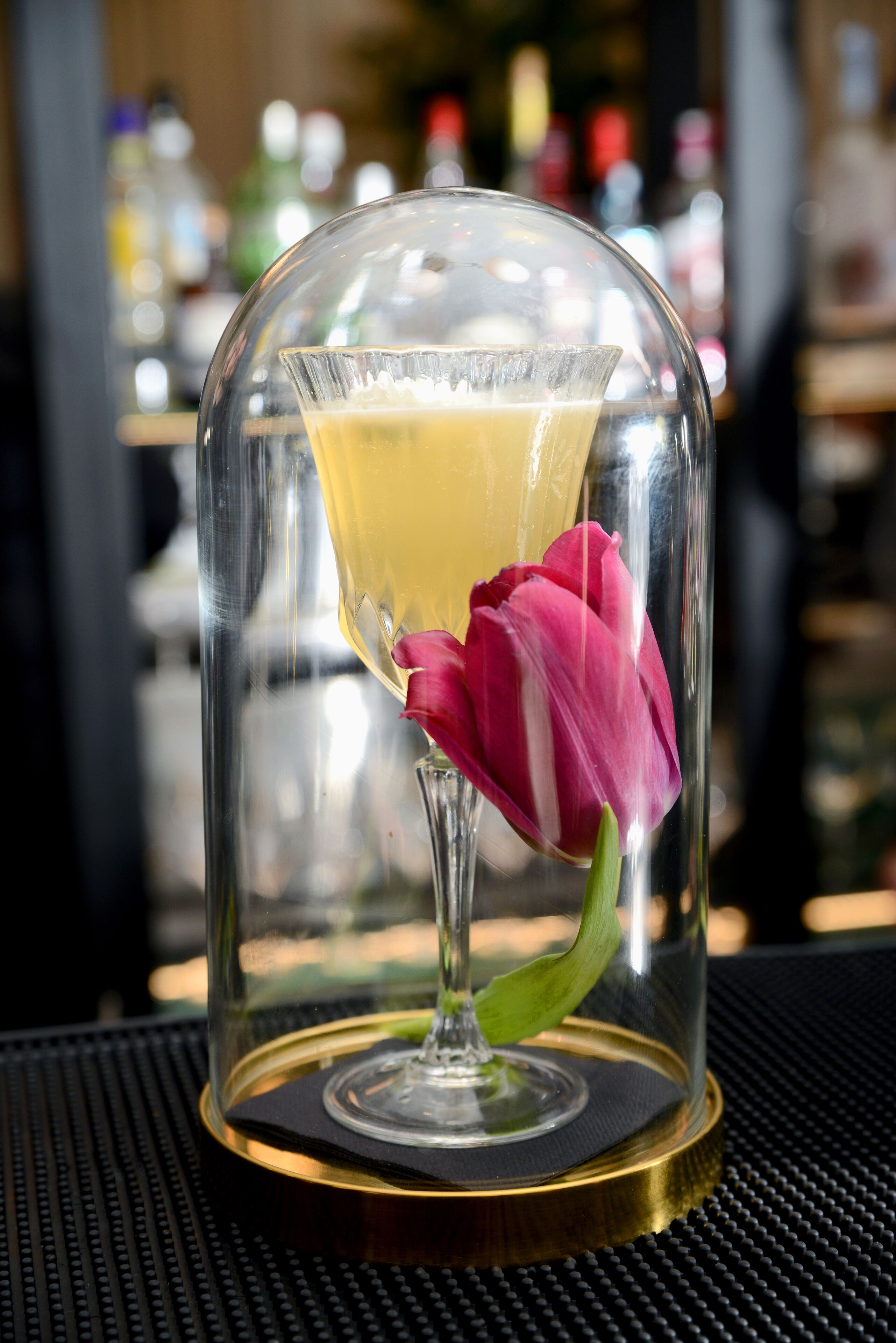 Een martini-glas met een geel drankje en een roze tulp in een glazen koepel op een tafel.