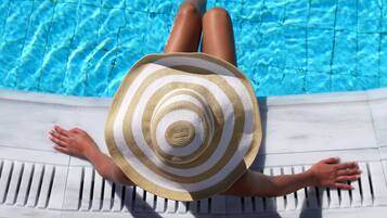 A person lying on a poolside lounge chair with a striped sun hat, facing a clear blue pool.