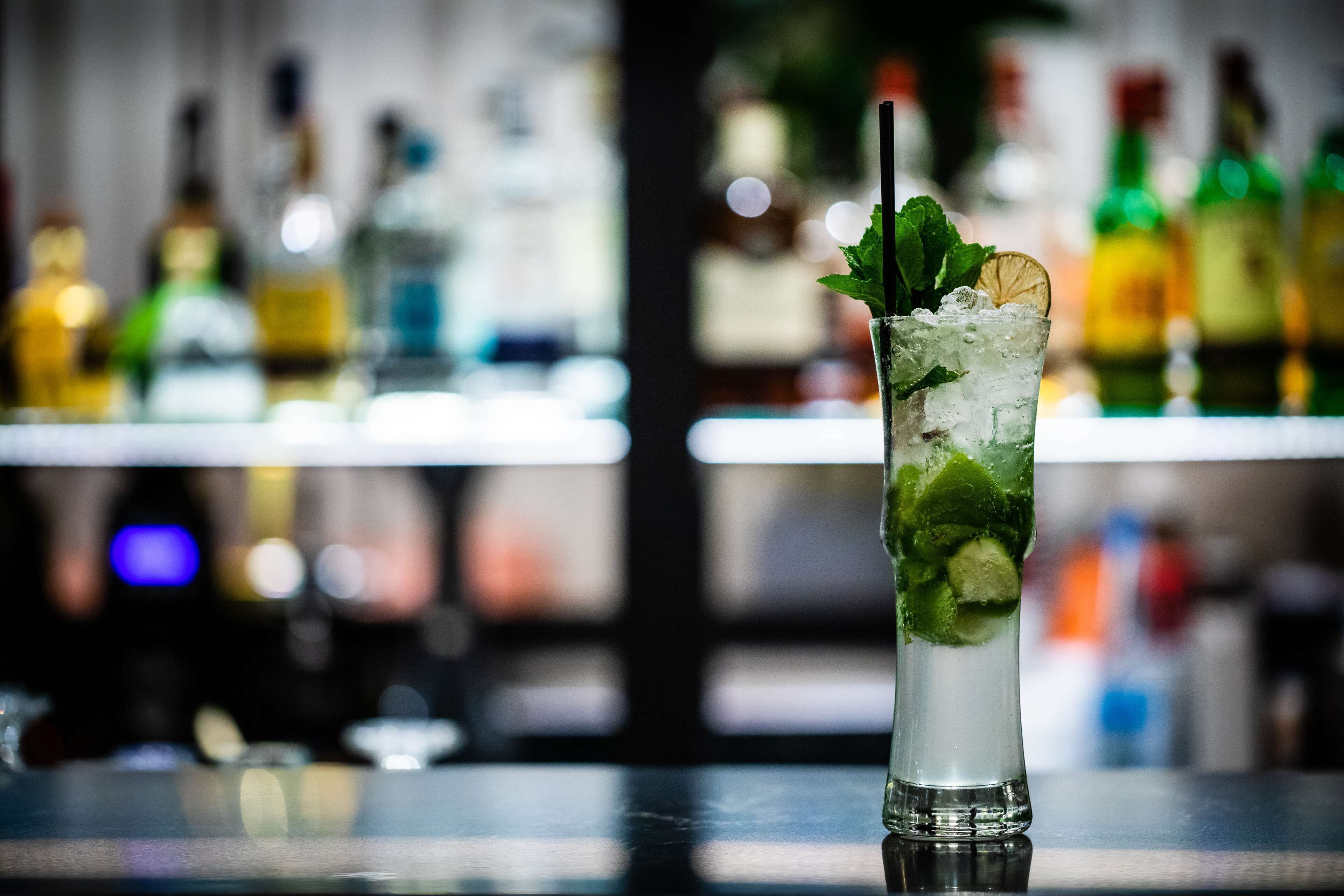 Un grand verre contenant une boisson décorée de feuilles de menthe et d’une tranche de citron, posé sur le comptoir d’un bar, avec des bouteilles floues en arrière-plan.