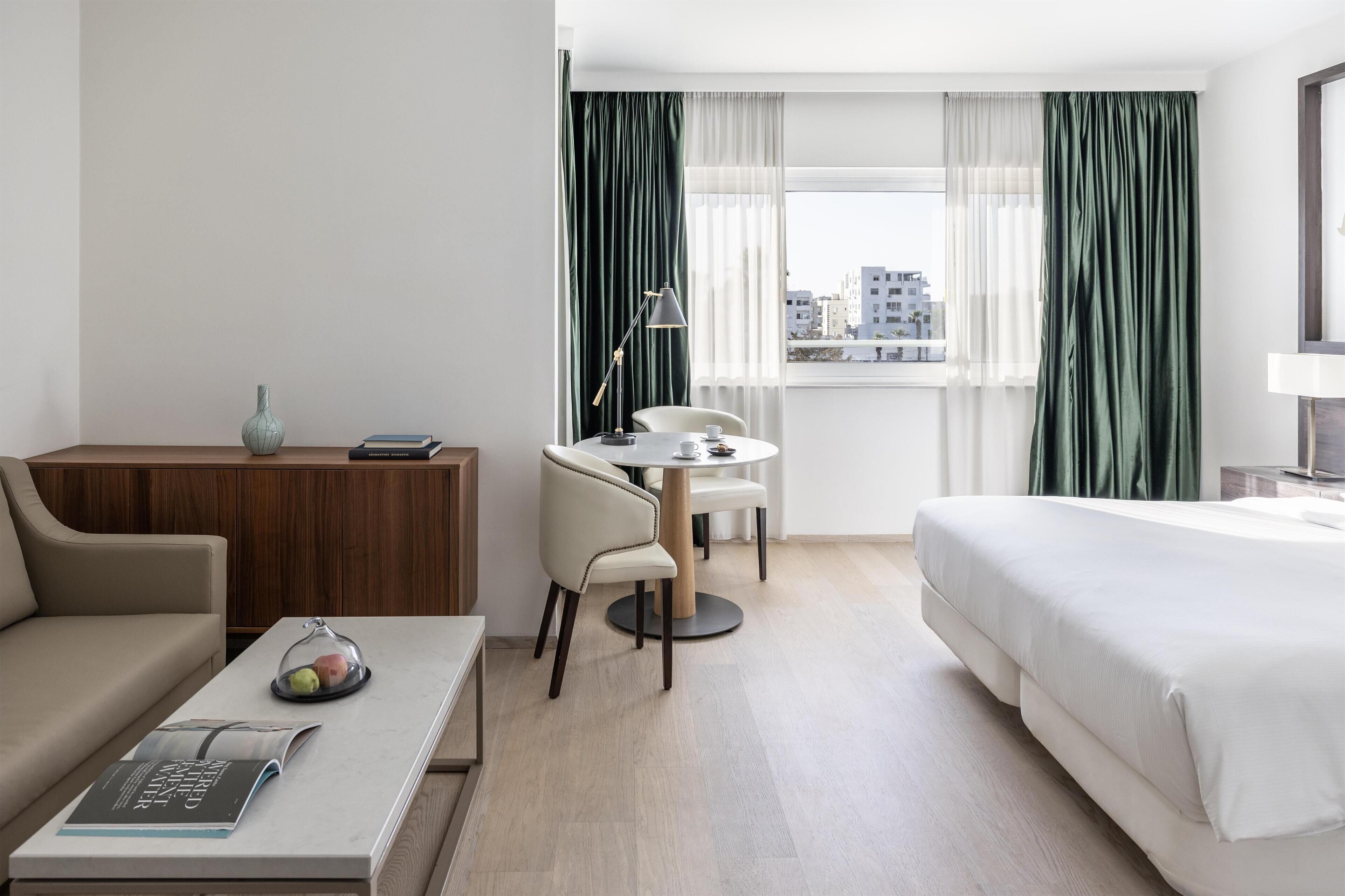 A hotel room with a bed, a desk, a chair, a small table, and a window with curtains.
