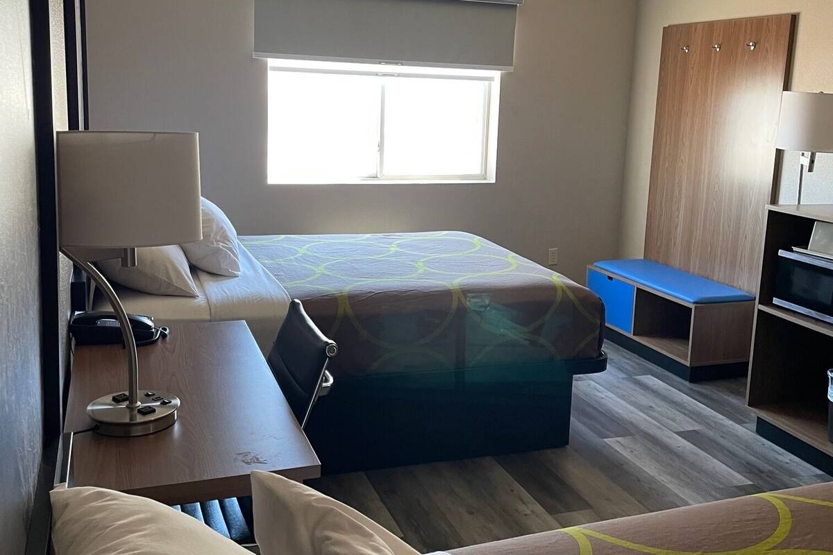A hotel room with two beds, a desk with a lamp, a wardrobe, and a window with blinds.