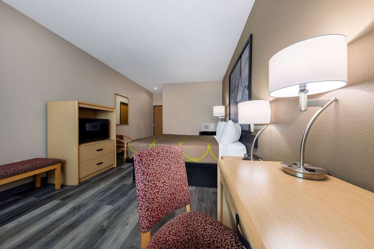 A hotel room with a wooden desk, a chair, a television, a bed, and a lamp.