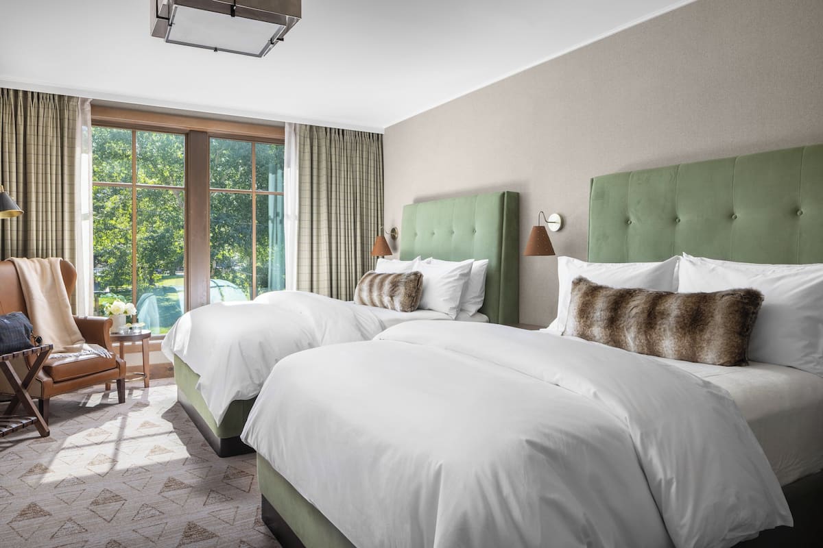 A hotel room with two large beds, a chair, a small table with a vase, and a view of trees through the window.