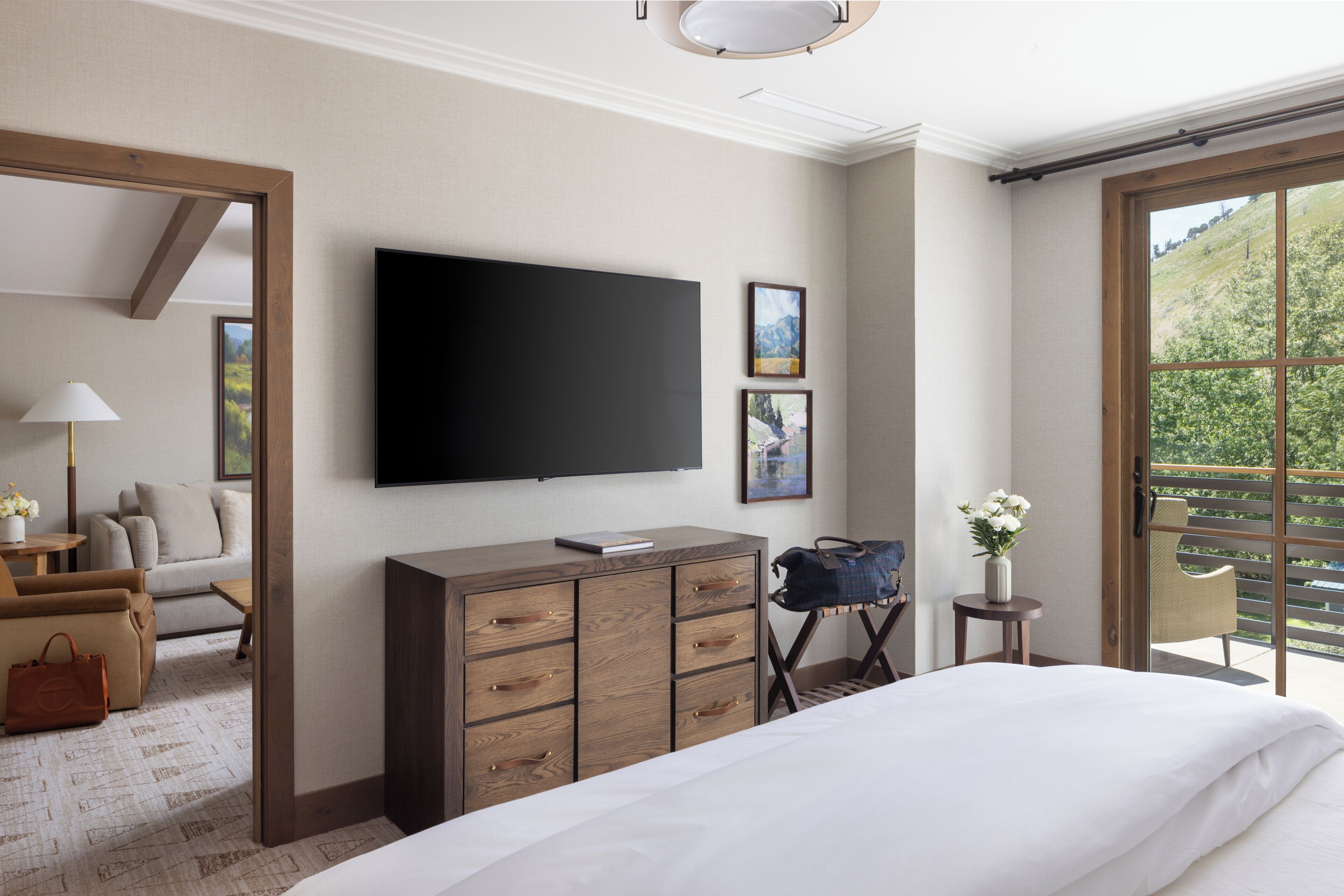 Et hotelværelse med en stor seng, et fladskærms-tv monteret på væggen, en kommode, en stol og et lille bord med en vase med blomster.