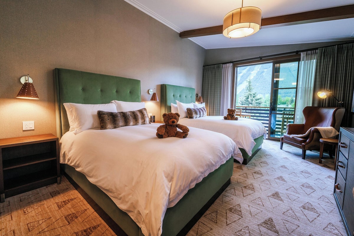 A hotel room with two beds, a large window with a mountain view, a chair, and a dresser.