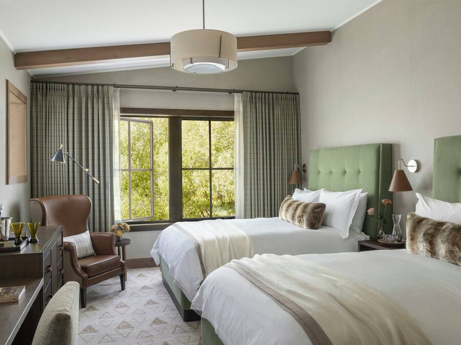 A hotel room with two beds, a desk, a chair, a lamp, and a window with curtains.