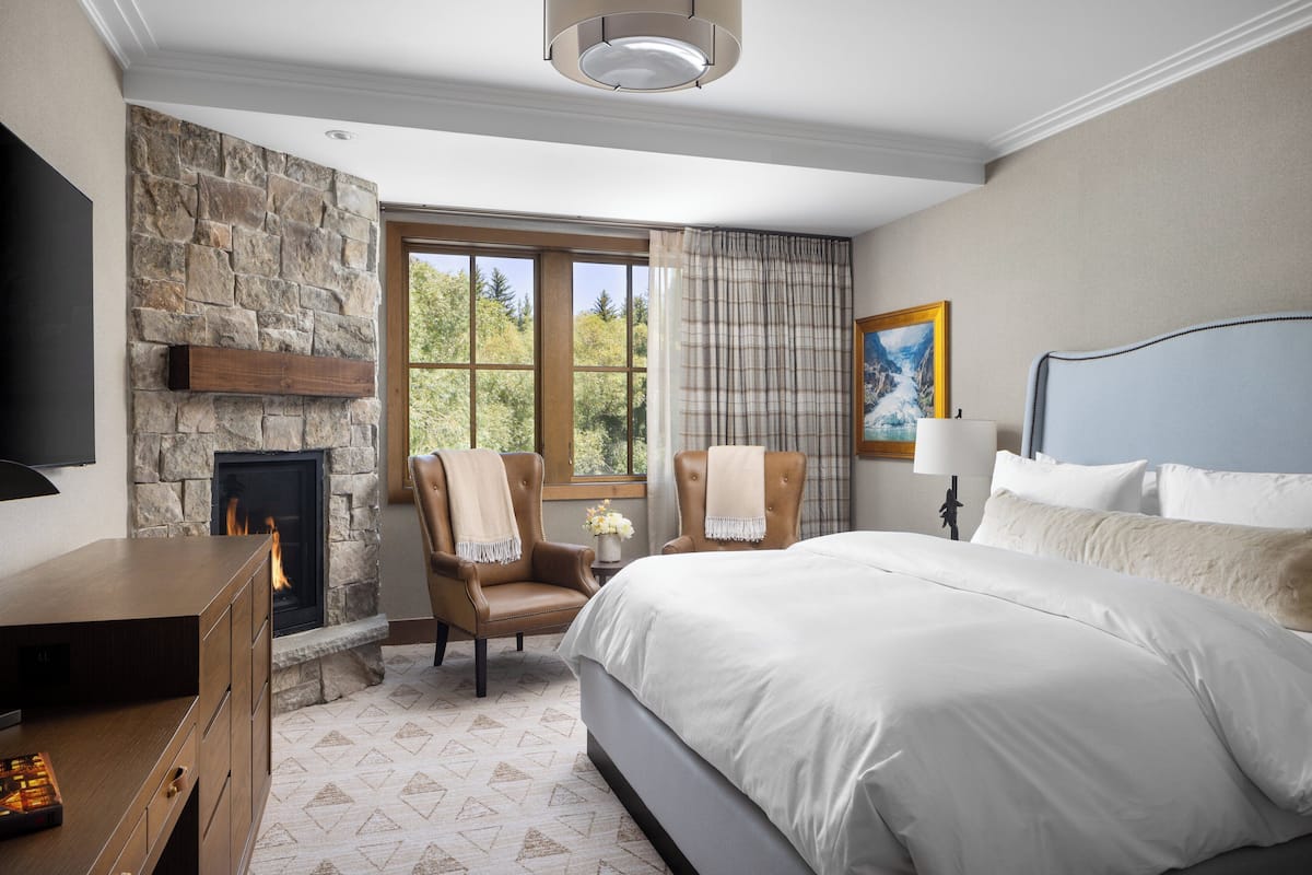 A bedroom with a stone fireplace, a bed with white bedding, a desk with a flat-screen TV, and a window with curtains.