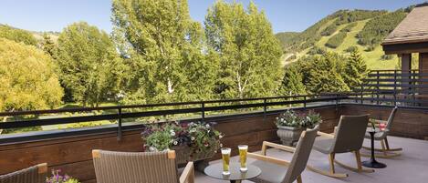 A terrace with wicker chairs and tables, overlooking a scenic landscape with trees and hills.