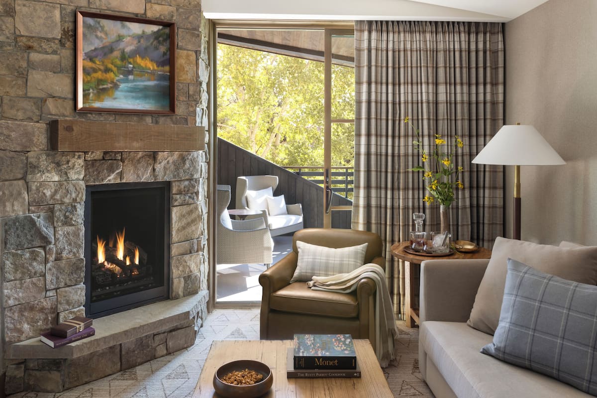 A living room with a stone fireplace, a sofa, a chair, a table with a book, and a view of a balcony.