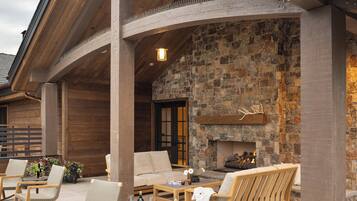 An outdoor patio area with wooden furniture, a stone fireplace, and a view of the interior.