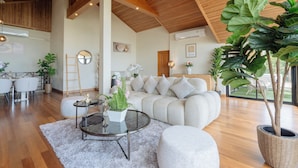 Ein geräumiges Wohnzimmer mit einer großen Couch, einem Couchtisch und einer Zimmerpflanze. Es gibt einen Essbereich mit Tisch und Stühlen, und im Hintergrund eine Küche.