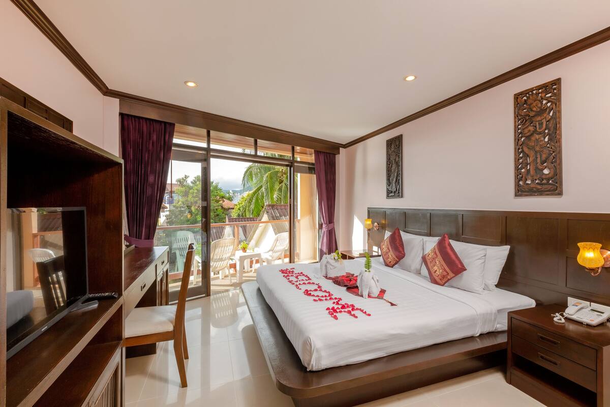 A hotel room with a large bed, a desk, a TV, and a balcony with chairs.