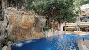 Ein Pool mit steiniger Kante, umgeben von viel Grün und einem Gebäude mit Außenbereich.