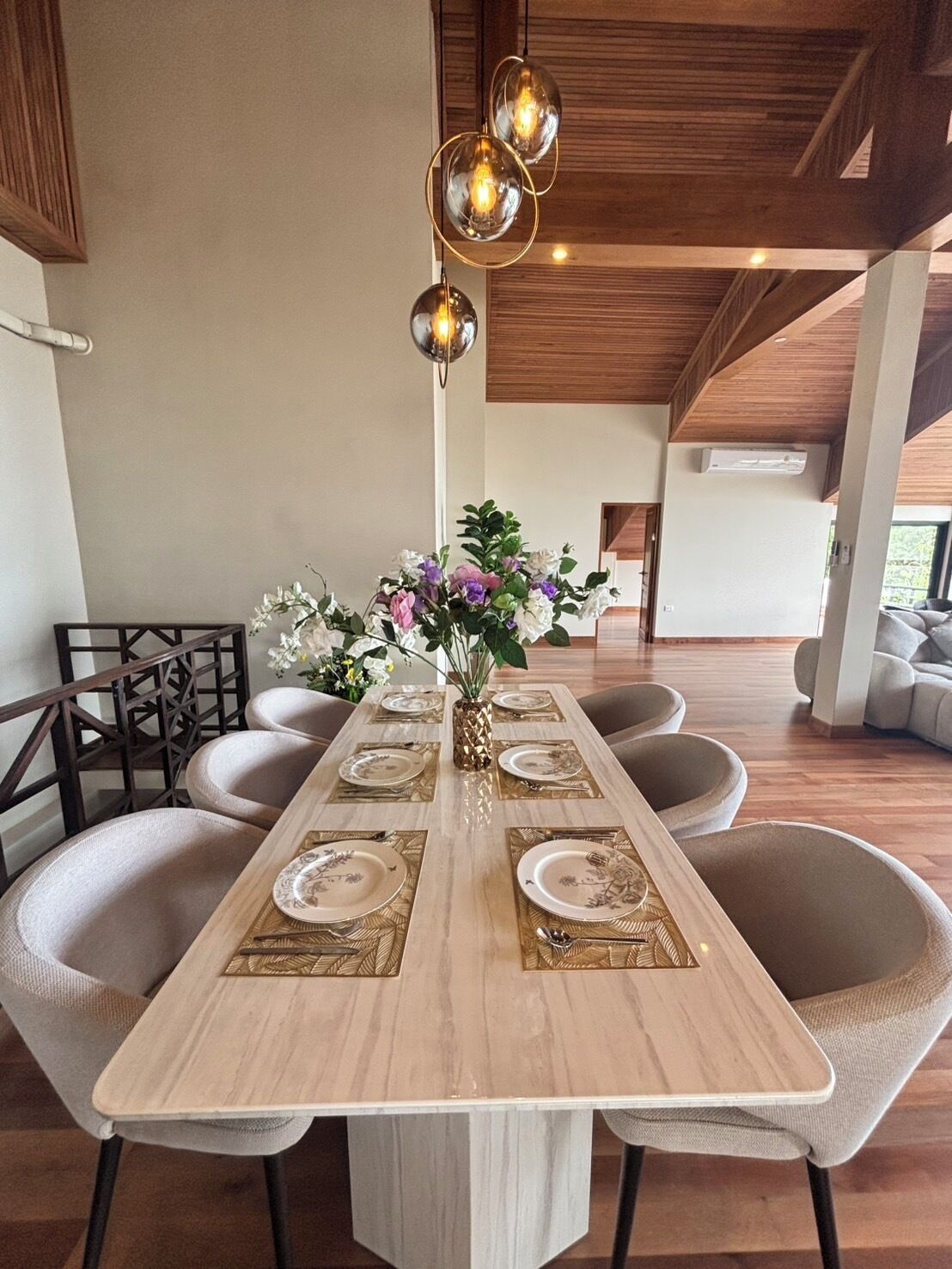 A dining area with a long table set for four, surrounded by cushioned chairs. A vase with flowers is placed in the center.