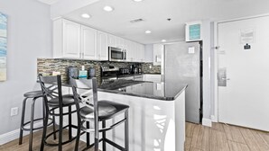 A modern kitchen with white cabinets, a central island with bar stools, a stainless steel refrigerator, and a microwave. - SunDestin Beach Resort (Destin)