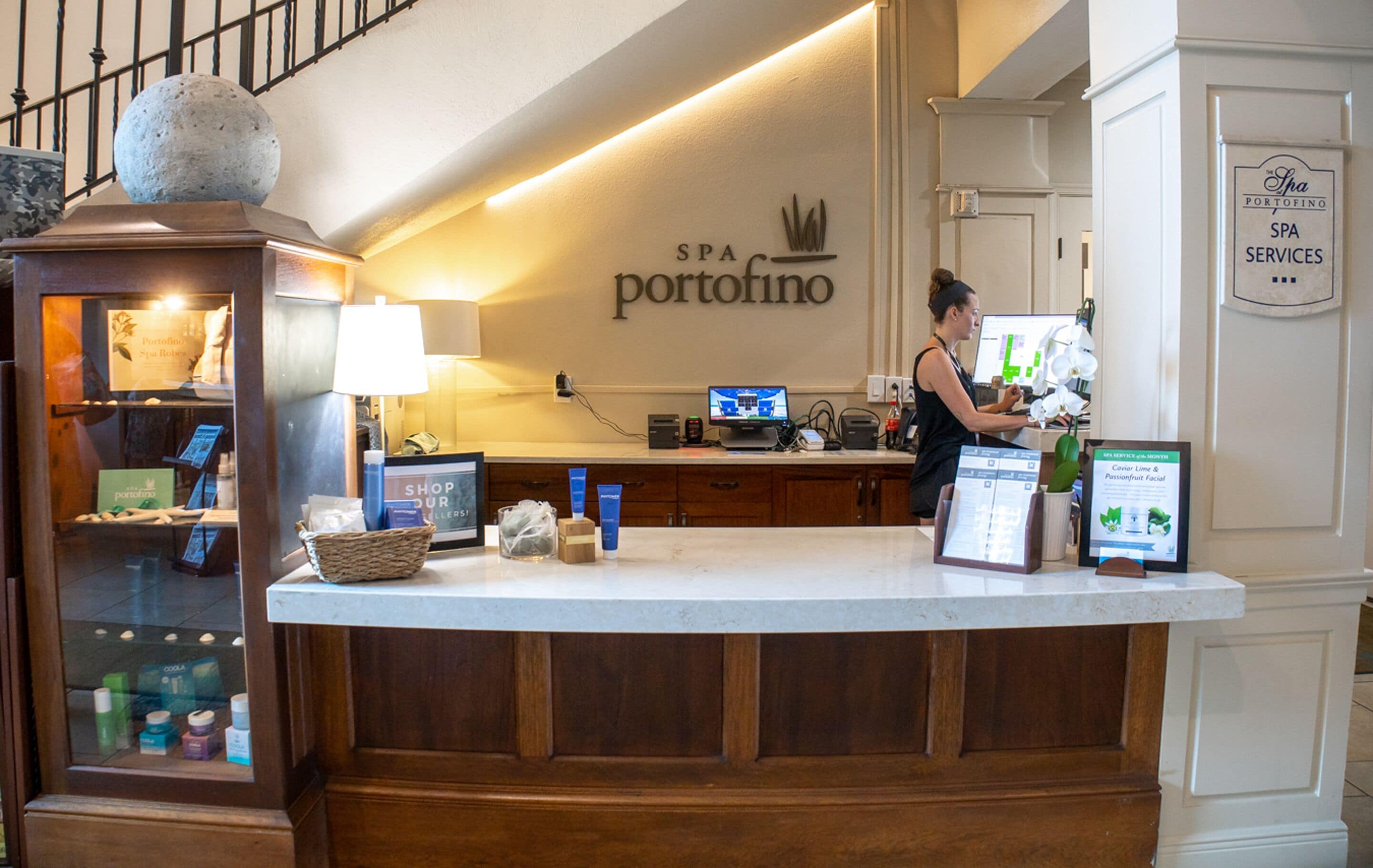 A reception desk with a computer, a woman standing behind it, and a sign that reads "Spa Portofino Spa Services".