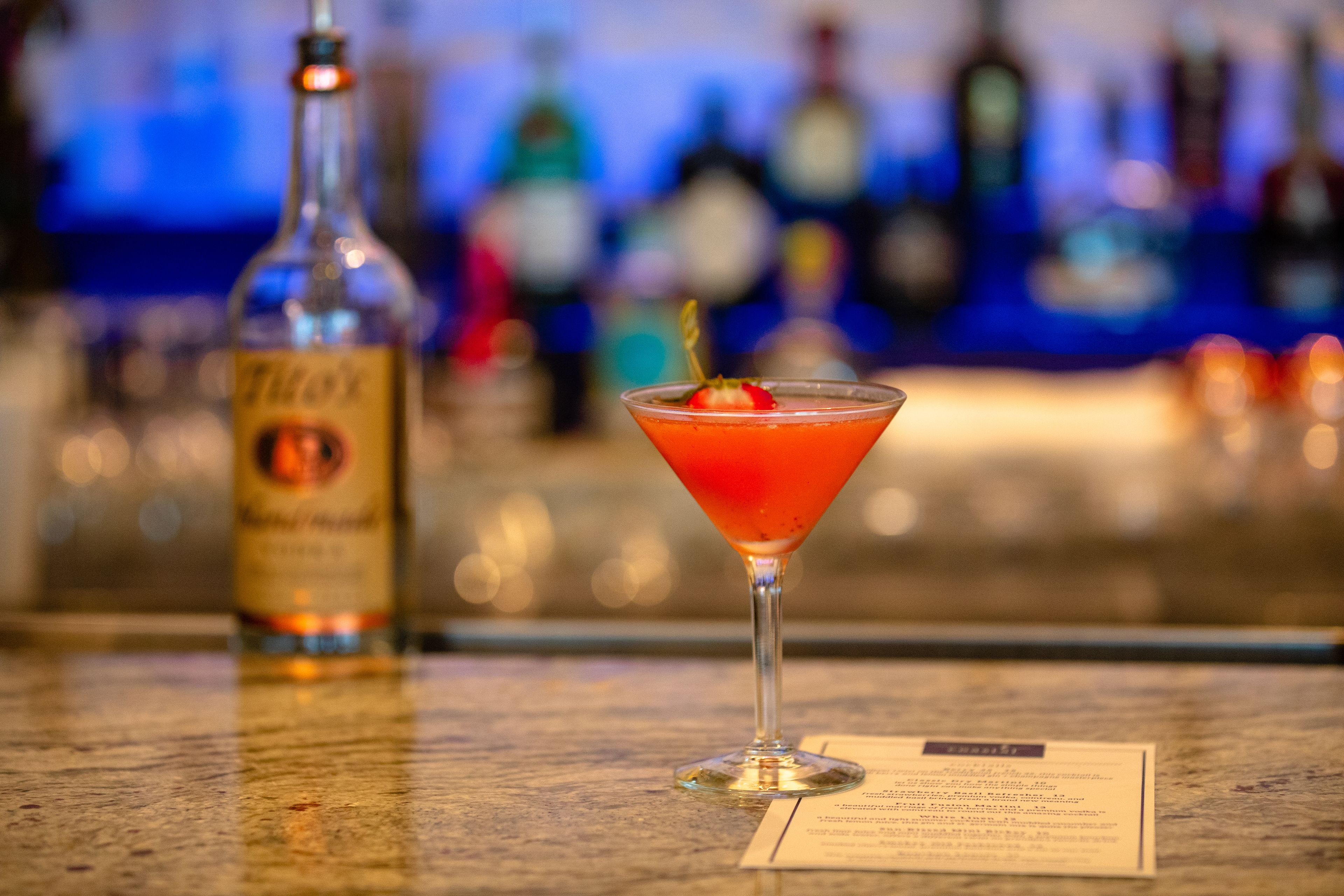 A martini glass with a garnish on a bar counter, a bottle of liquor, and a menu.